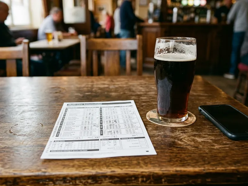 A betting slip showing a multi-leg accumulator beside a phone and a pint on a pub table